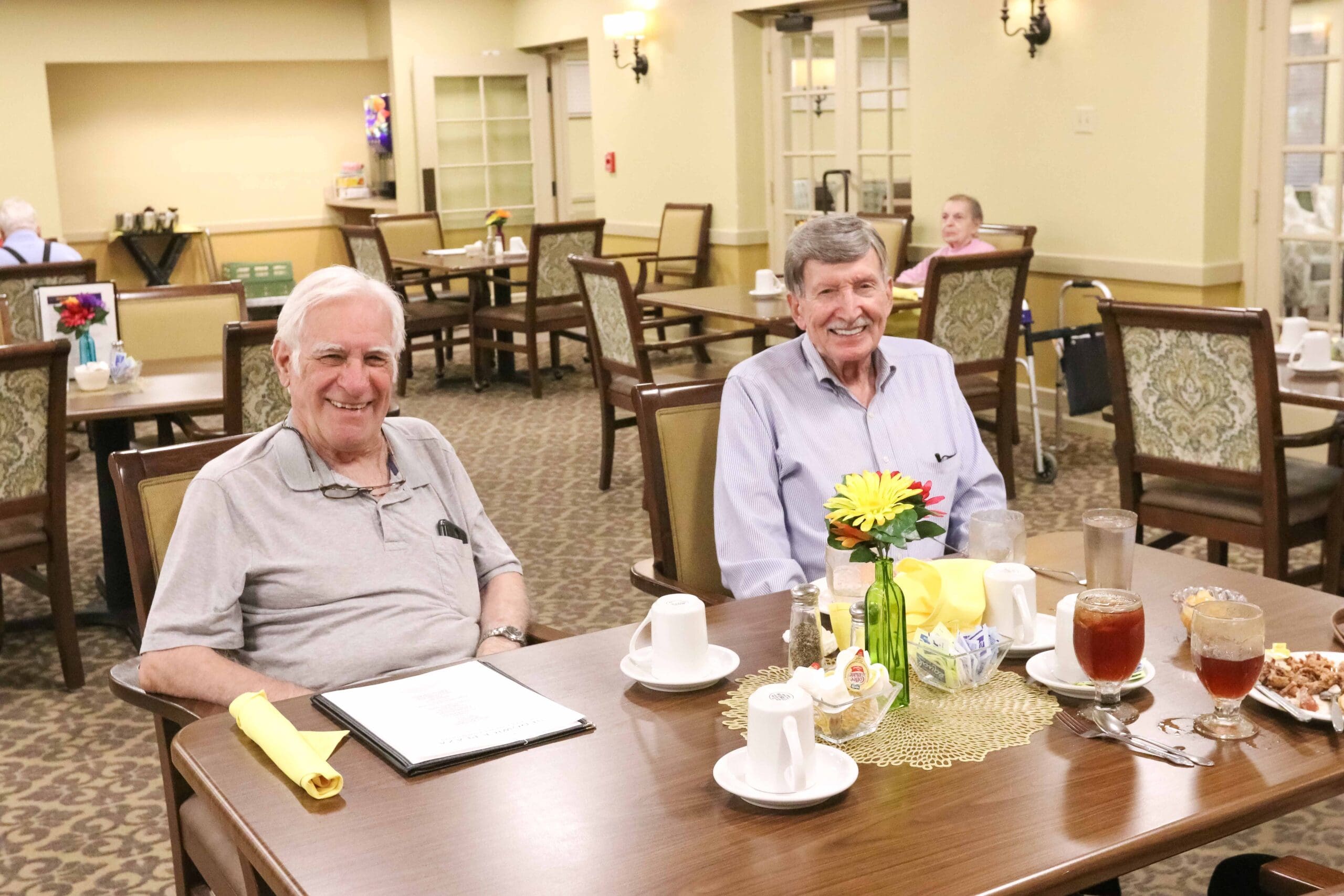 Two male residents sit together in the dining room enjoying dinner and drinks together at Inspirations of Bartlesville.